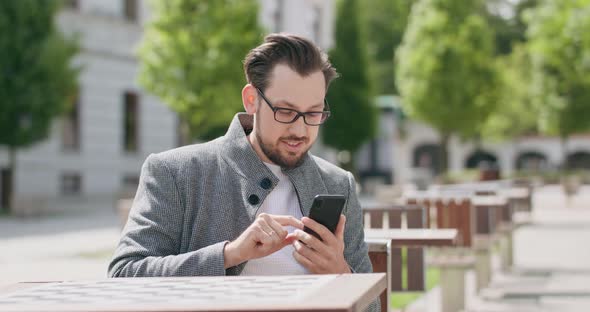 Young Man Wearing Glasses with Mustaches and a Beard is Sitting at the Table in the Square is