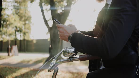 Man Wearing Suit Looks at the Phone Leaning on the Bike in the Park Close Shot