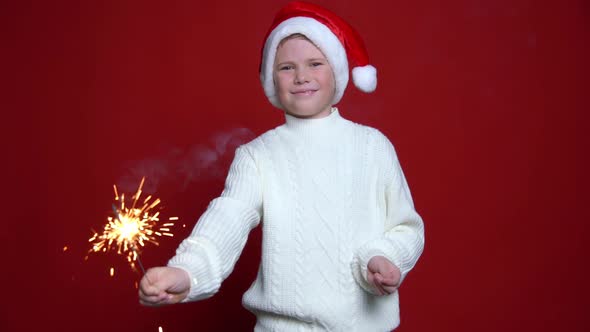 Happy Little Boy in Santa Claus Cap Holding Bengal Light on the Red Background