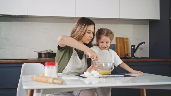 Woman with Daughter in Aprons Whisks Egg in Bowl at Table