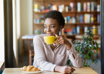 Happy black lady with cup of coffee and croissant enjoying peaceful morning, looking out window at / Photodune, Misc