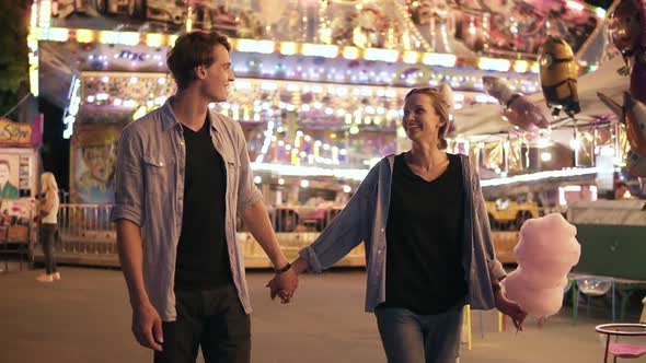 Beautiful Young Couple Visiting Funfair Smiling Holding Hands Walking and Eating Cotton Candy Night