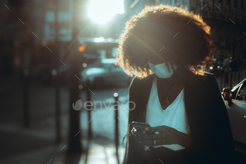  protective mask phoning on a city street on a sunny evening; a black female with a curly afro hair in an anti-virus mask using a smartphone outdoors