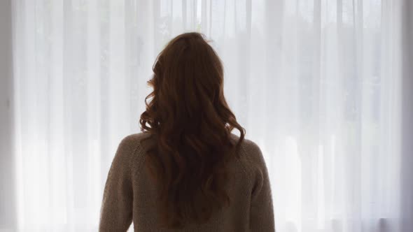 Rear view of woman drawing curtains of the window at home