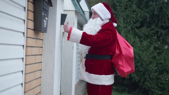 Side View Santa Ringing Doorbell Standing Outdoors with Red Gifts Bag