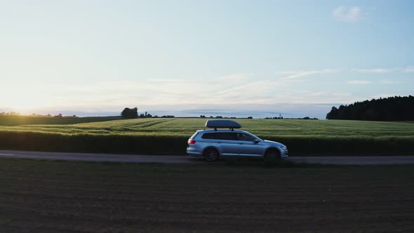 A Car with Roof Rack Drives Along a Road Through Green Meadows and Arable Fields