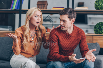 emotional upset couple sitting on couch and quarreling about smartphone, mistrust concept