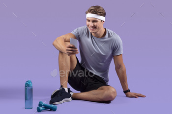 Full length portrait of sporty millennial guy using smartphone while resting after workout on lilac / Photodune, Misc