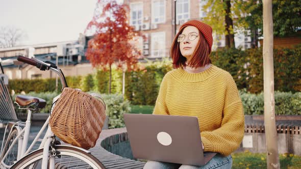 Young Hipster Woman Sitting on Bench in City Park Typing on Notebook Smiling