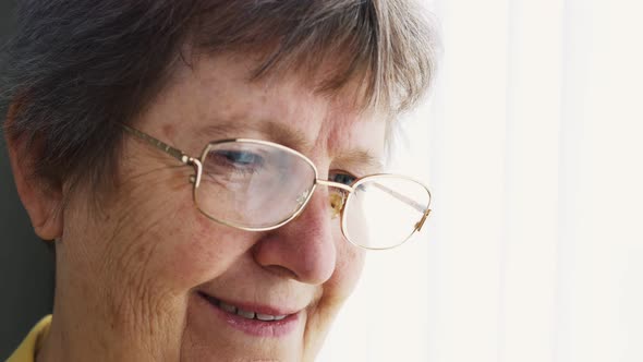 An Old Woman with Glasses Stands By the Window Her Face is in Marches and Her Head is Covered with