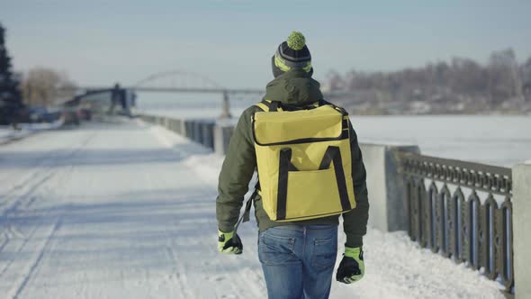 Back View of Courier with Large Yellow Thermo Bag Walking Along Snow Covered City Street