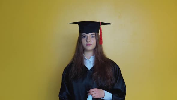 Caucasian Femal Student in a Black Gown and Hat on a Yellow Solid Background Looks at the Camera and