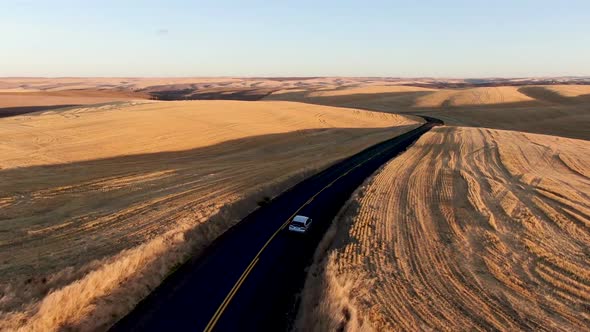 Drone follow shot of a car driving along a narrow country road among golden fields at sunrise or sun