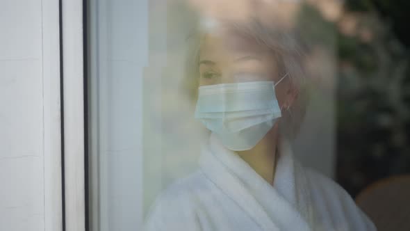 Shooting Through Window Sad Mature Woman in Covid19 Face Mask Standing Indoors Looking Out