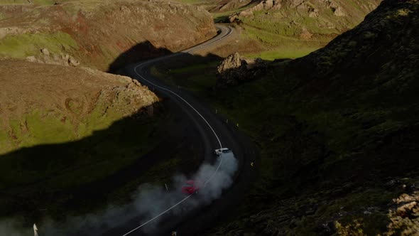 Cars Emitting Smoke While Drifting On Asphalt Road Through Green Rocky Hills. aerial tilt down