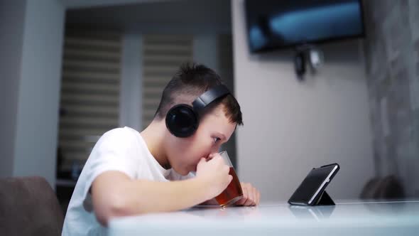 Boy Using Tablet At Home. Boy sitting on table and drinking tea while looking on his tablet