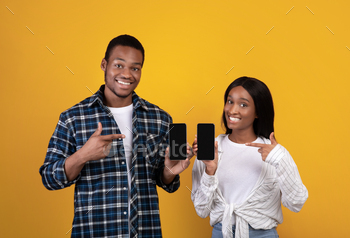  Smiling zoomers african american couple showing smartphones with blank screens and pointing fingers at gadgets, isolated on yellow background