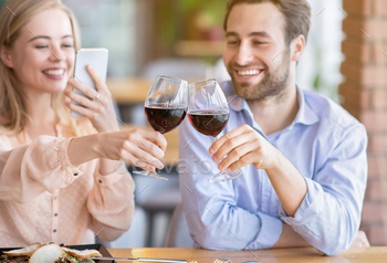 Young woman making photo of romantic toast with clinking wine glasses during festive dinner at cafe / Photodune, Misc