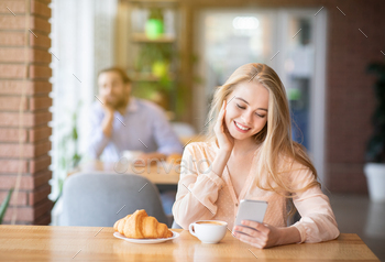 Beautiful young woman using smartphone during breakfast at cafe, young guy looking at her, empty / Photodune, Misc