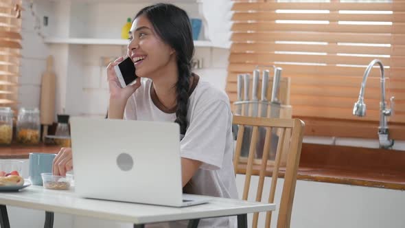 Young Asian woman happy smiling talking on the phone and using laptop in kitchen at home.