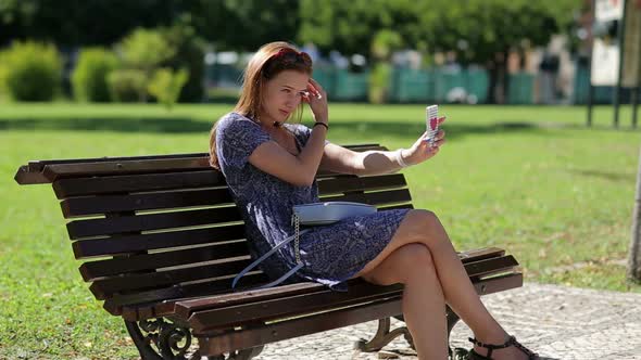 Young Beautiful Woman Make Selfy Holding Mobile Phone in Hand and Sitting on the Bench in the Park