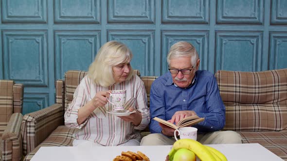 Retired Senior Couple Talking Drinking Tea Reading Book in Modern Living Home Room Lounge Together