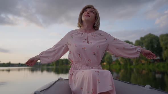 Medium Shot of Joyful Relaxed Queer Person in Pink Dress Stretching Hands Sitting in Boat on Summer