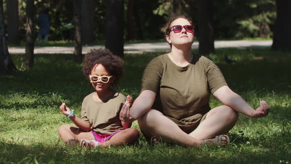 White Woman and Her Black Daughter Sitting on the Grass in the Park and Meditating