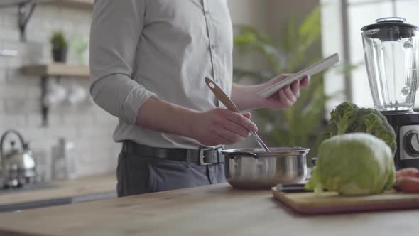 Unrecognizable Man in the Shirt Cooking Soup Checking Recipe on the Tablet in the Kitchen at Home