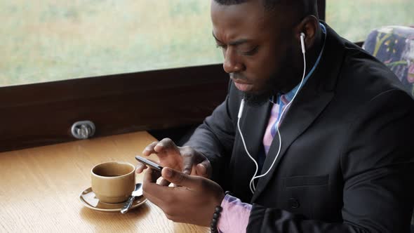 Black Man is Listening Music in Earphones Browsing Smartphone Sitting in Cafe