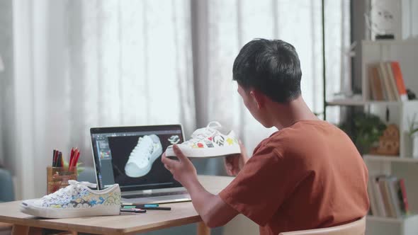 Asian Boy Footwear Designer Holding And Looking At Sneakers While Designing Shoe On A Laptop At Home