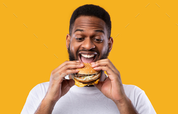 ood Standing Over Yellow Studio Background. African Man Enjoying Hamburger. Unhealthy Nutrition And Binge Eating Habit