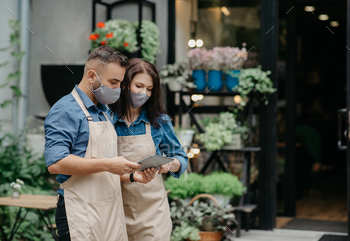  millennial man and female in aprons and protective masks use digital tablet and blog and make advertisement or marketing studio near flower shop