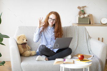 enager girl in glasses showing ok sign gesture sitting on the sofa with crossed legs and using laptop, doing homework, recommending online school