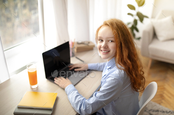 e girl sitting at desk and using laptop computer, doing homework, browsing internet, watching video or movie, looking back at camera