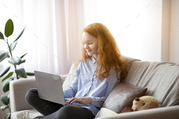 haired teenage girl sitting on the sofa and using laptop computer, doing homework, browsing internet, watching video or movie, typing on keyboard