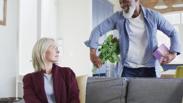 Happy senior diverse couple in living room sitting on sofa, giving flowers and present