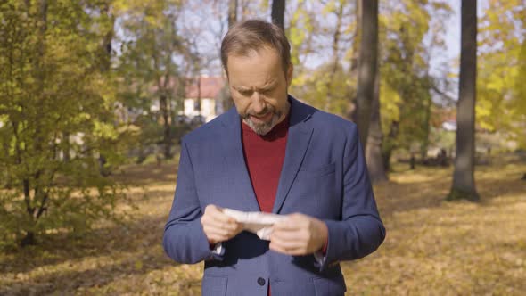 A Middleaged Handsome Caucasian Man Blows His Nose in a Park in Fall  Trees in the Background