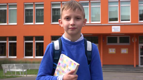 A Young Boy Holds a Book and Looks at the Camera in Front of an Elementary School