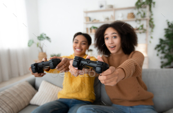 tion and rest at home. Smiling surprised excited millennial african american sisters have fun with joysticks in living room interior, free space