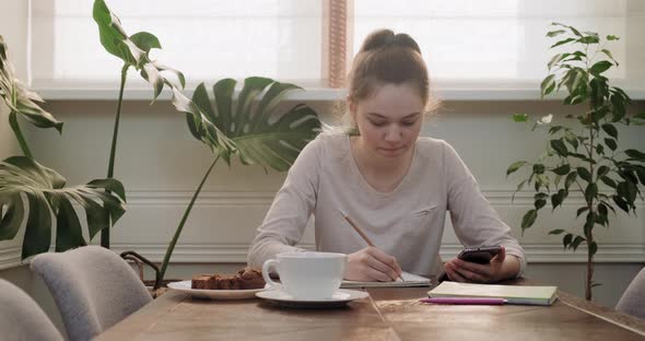 Teen Girl Sitting at Home at Table Writing Lessons Using Smartphone for Learning