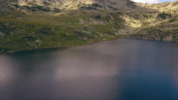 Wide aerial drone slowly flying over a lake towards a green shore line leading up to a mountain rang