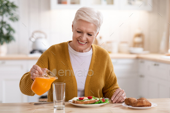 ing fresh juice into glass in kitchen, having lunch alone, copy space. Happy grey-haired senior woman eating healthy food, home interior