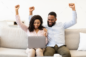 n and woman happy with money win, satisfied about success. Black couple shaking fists and looking at laptop computer, sitting on the couch at home