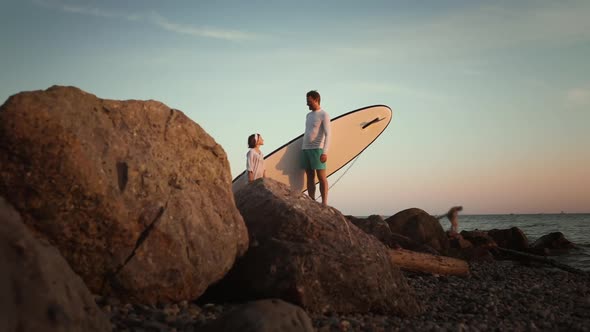 Man and preschool girl standing on the beach with sup board. Ocean's surface on the background