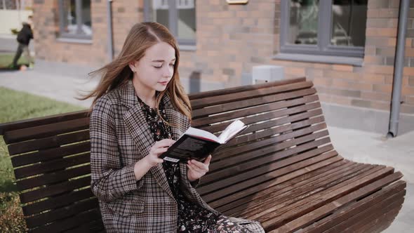 A young girl with blonde hair reads a book on a bench