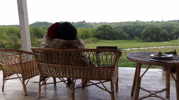 A Couple in Love is Sitting on Terrace on Cane Bench Hugging and Covering in Blanket