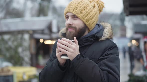 Portrait of Young Bearded Caucasian Man in Funny Yellow Hat and Winter Coat Warming Up with Hot