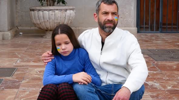 Father and Daughter with Blue and Yellow Ukrainian Flag Painted on Cheeks Sitting on the Street