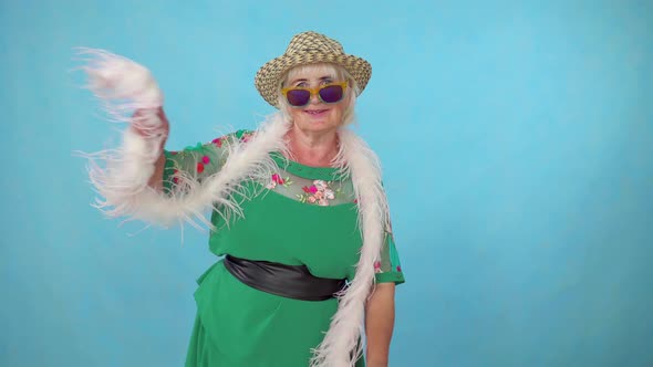 Cheerful Expressive Energetic Old Woman in a Hat and Boa Dancing on a Blue Background